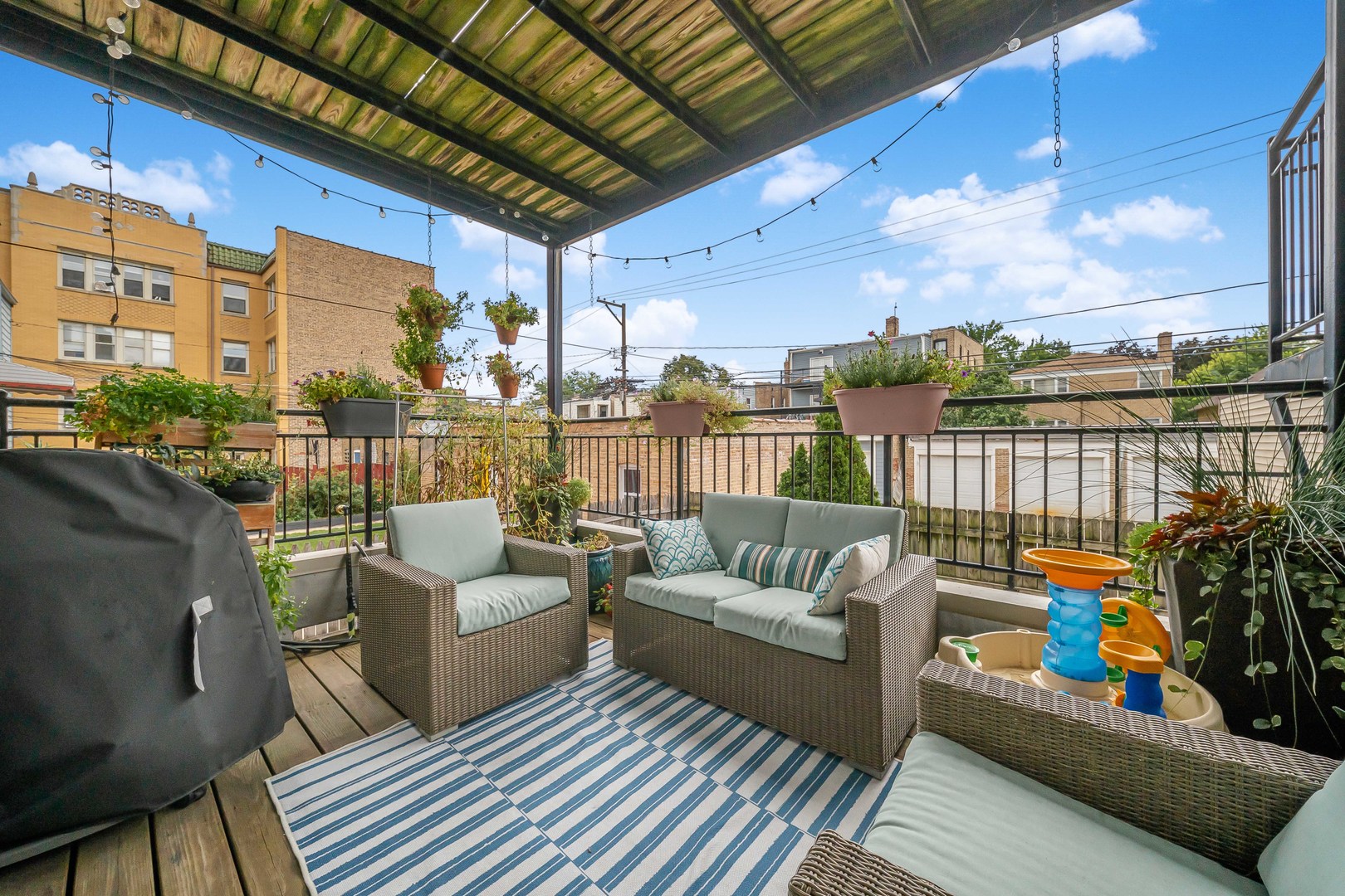 Furnished deck with pergola, wicker seating, potted plants, and a striped rug overlooking neighboring buildings.