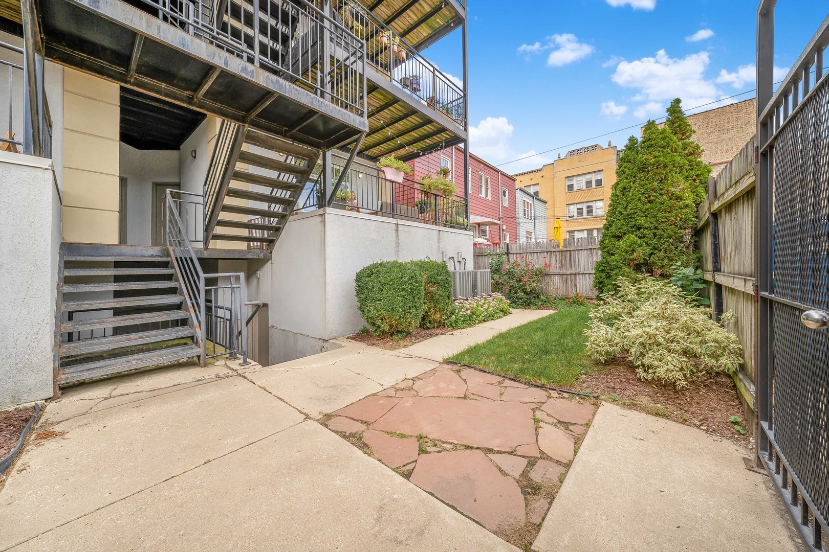 Landscaped courtyard with a stone patio, fenced lawn, flower beds, and metal exterior stairs.