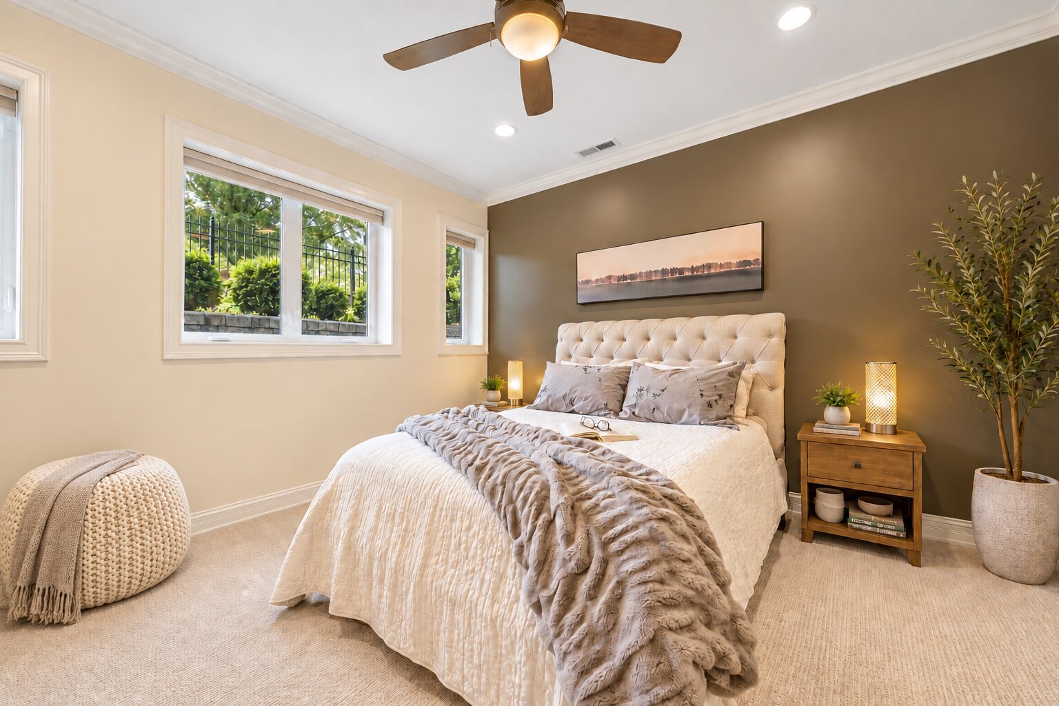 Bedroom with a cream tufted bed, dark accent wall, and large windows overlooking greenery.