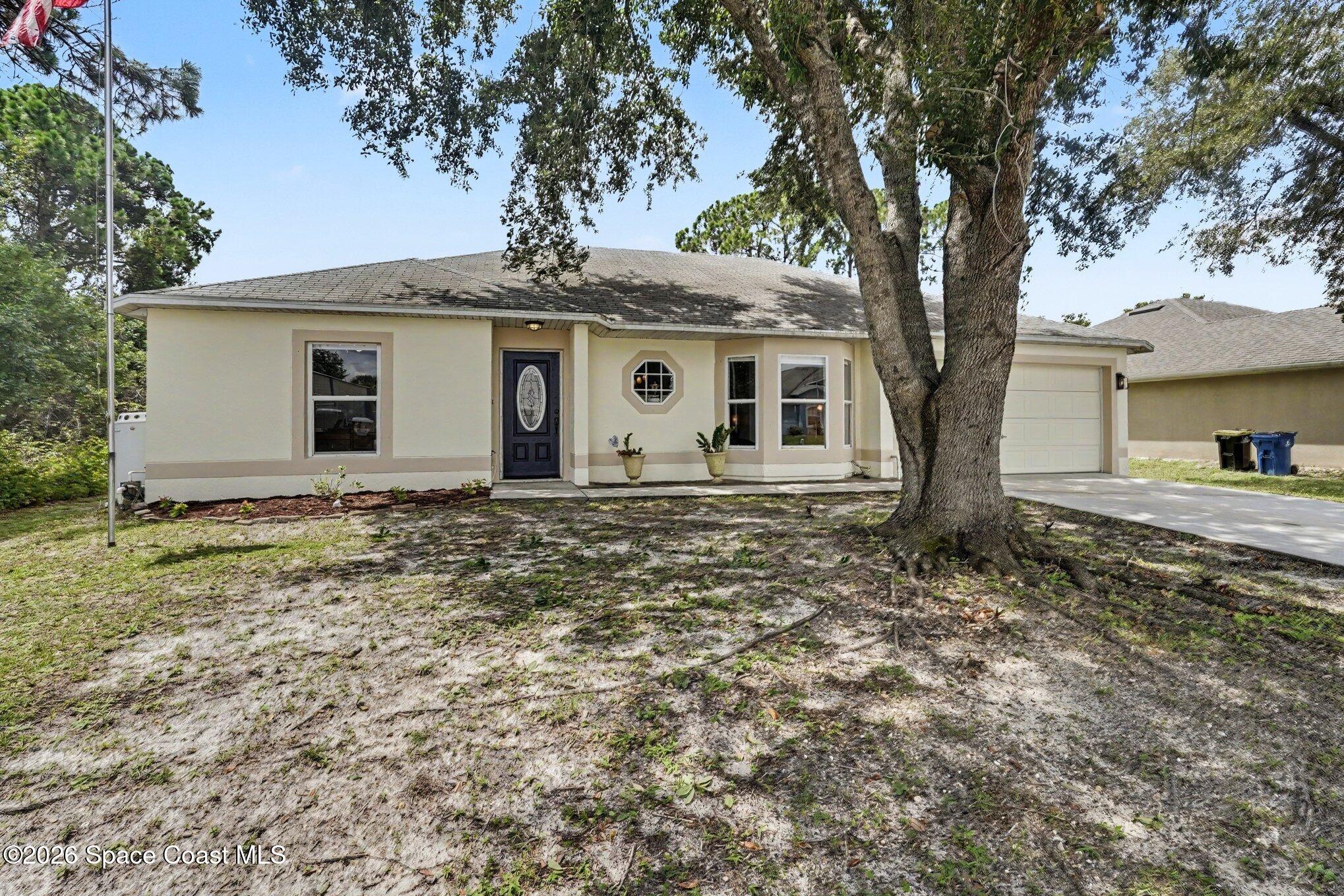 Single-story cream house with an attached garage and a large shade tree in the sandy front yard.