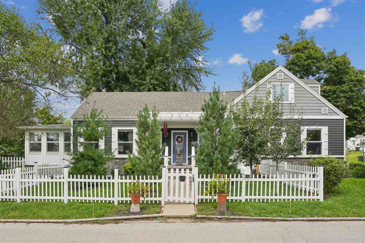 Gray gabled house with a covered porch behind a white picket fence and mature trees.
