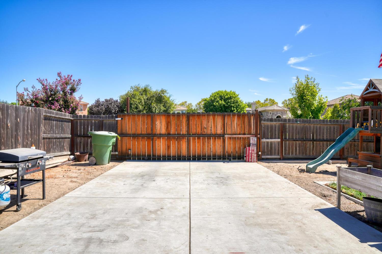 Backyard patio with a wide concrete slab, wood fencing, a grill, and a play structure with a slide.