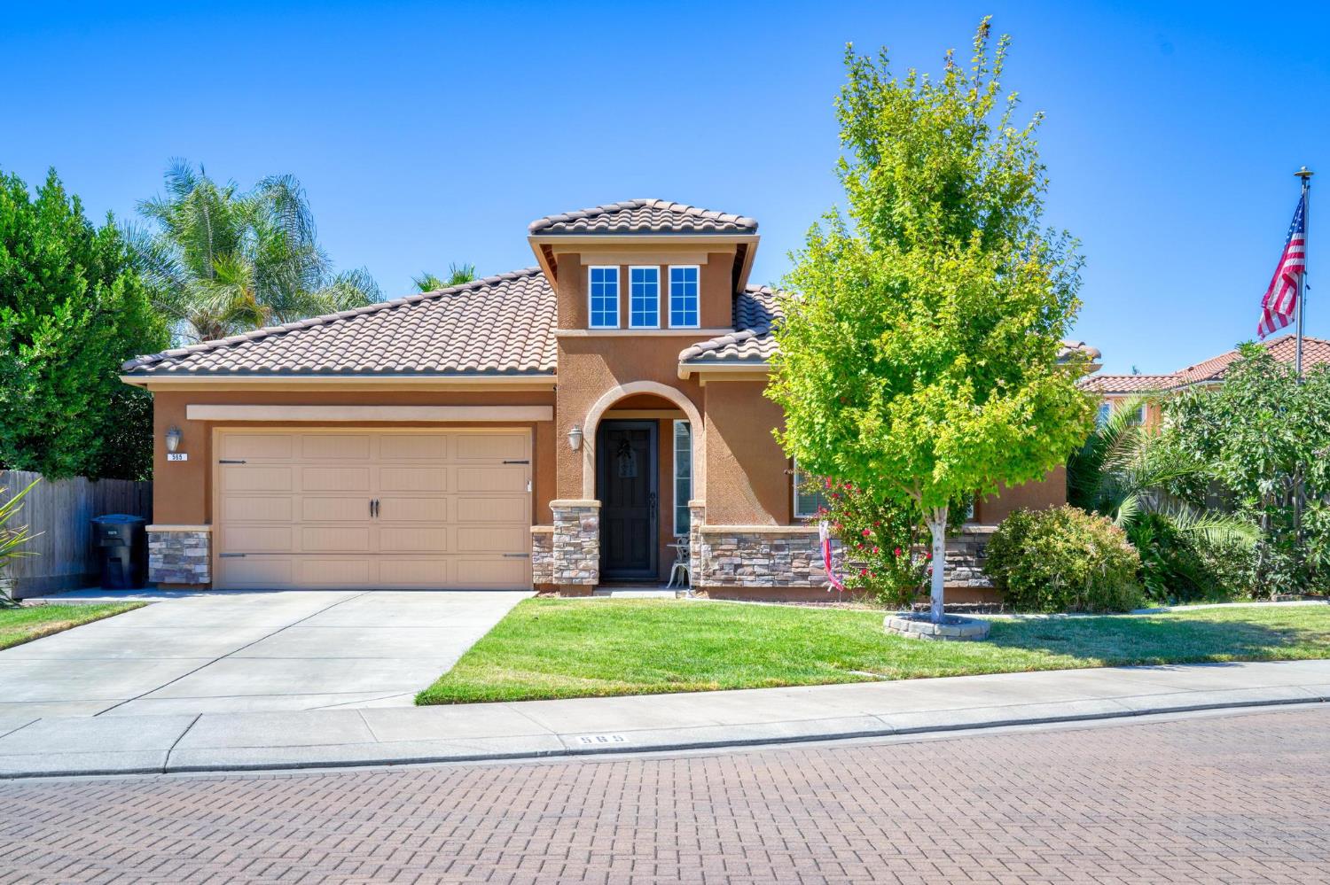 Tan stucco home with a tiled roof, arched entry, two-car garage, and leafy front-yard tree.