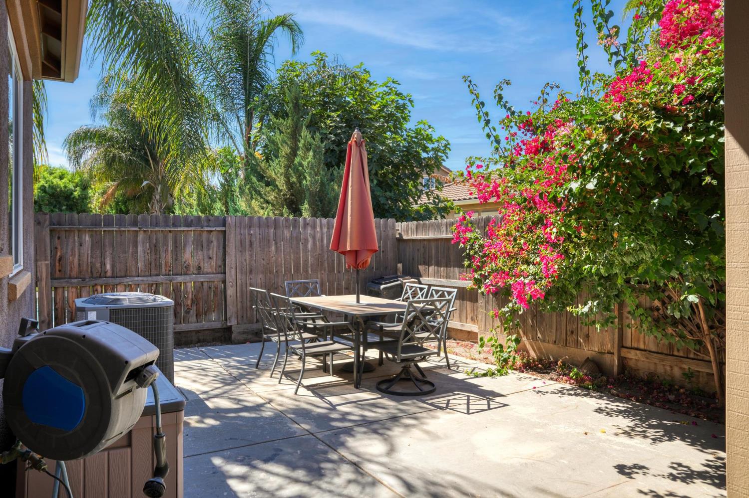 Backyard patio with outdoor dining set, closed umbrella, wooden fence, and flowering bougainvillea.