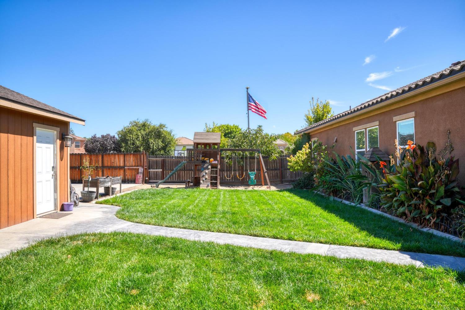 Fenced backyard with a broad lawn, wooden playset, shed, and American flag.