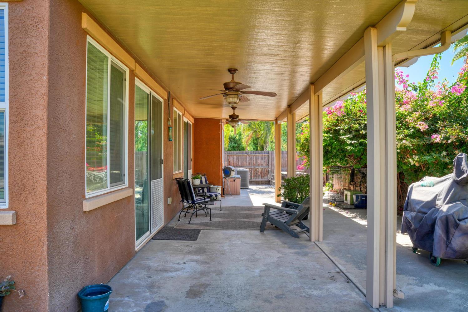 Covered backyard patio with ceiling fans, outdoor chairs, and flowering shrubs beside the home.
