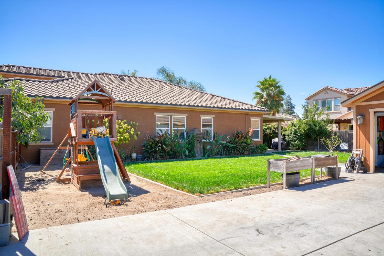 Backyard with a green lawn, wooden playset and slide, raised garden beds, and concrete patio.