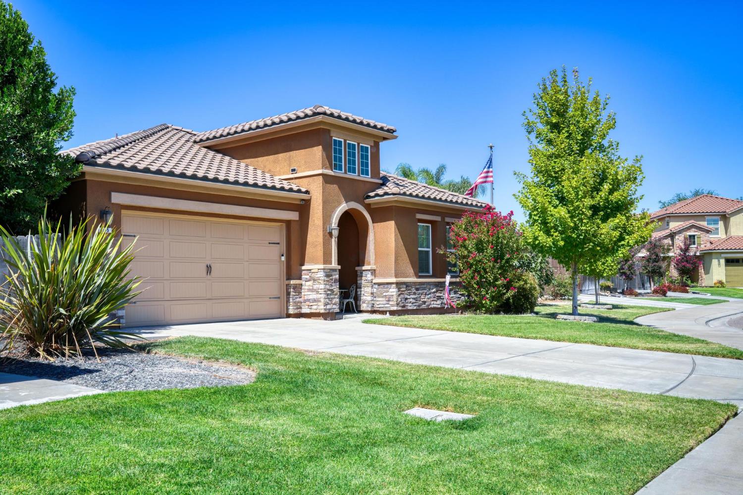 Two-story tan stucco home with tiled roof, arched entry, stone accents, and landscaped front yard.
