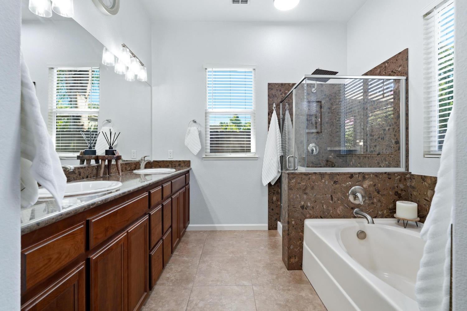 Bright bathroom with a double wood vanity, glass shower, stone tile, and bathtub.