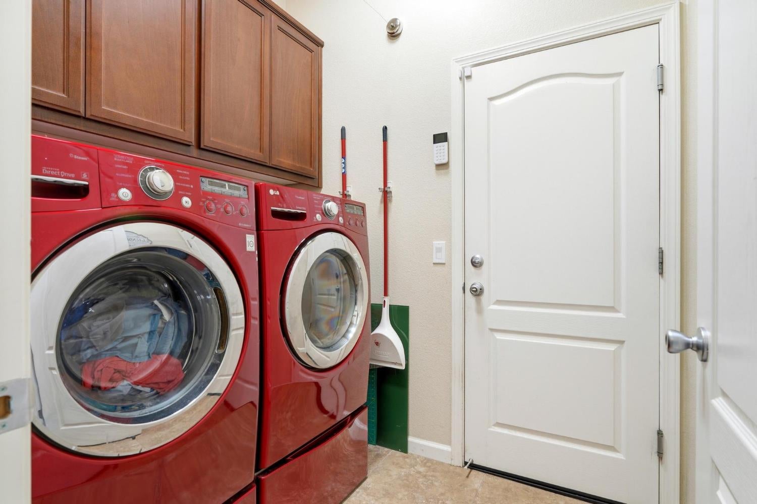Laundry room with red front-loading washer and dryer beneath dark wood cabinets, beside a white paneled door.