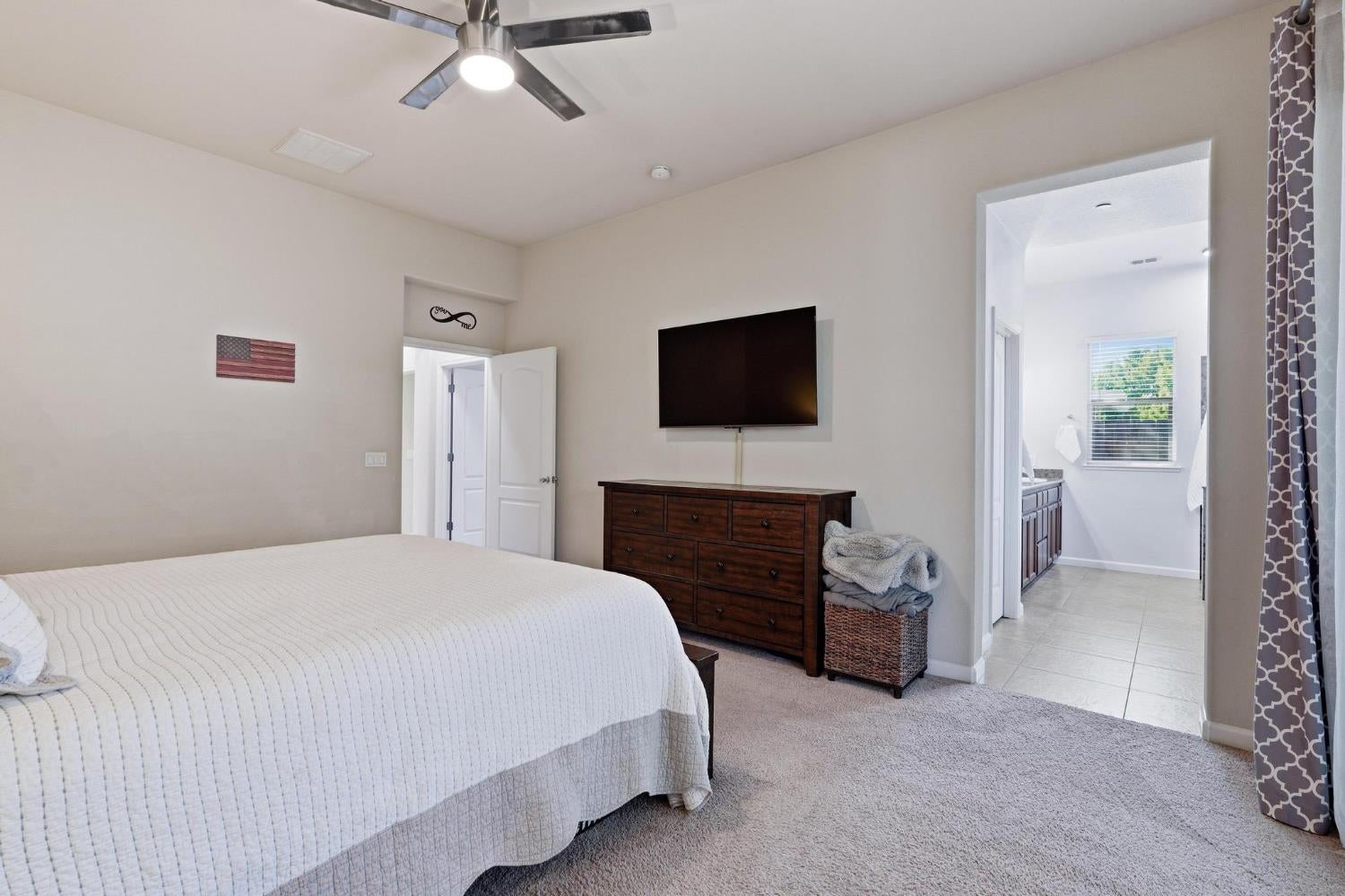 Bedroom with a white-covered bed, dark wood dresser, wall-mounted television, and open ensuite bathroom.