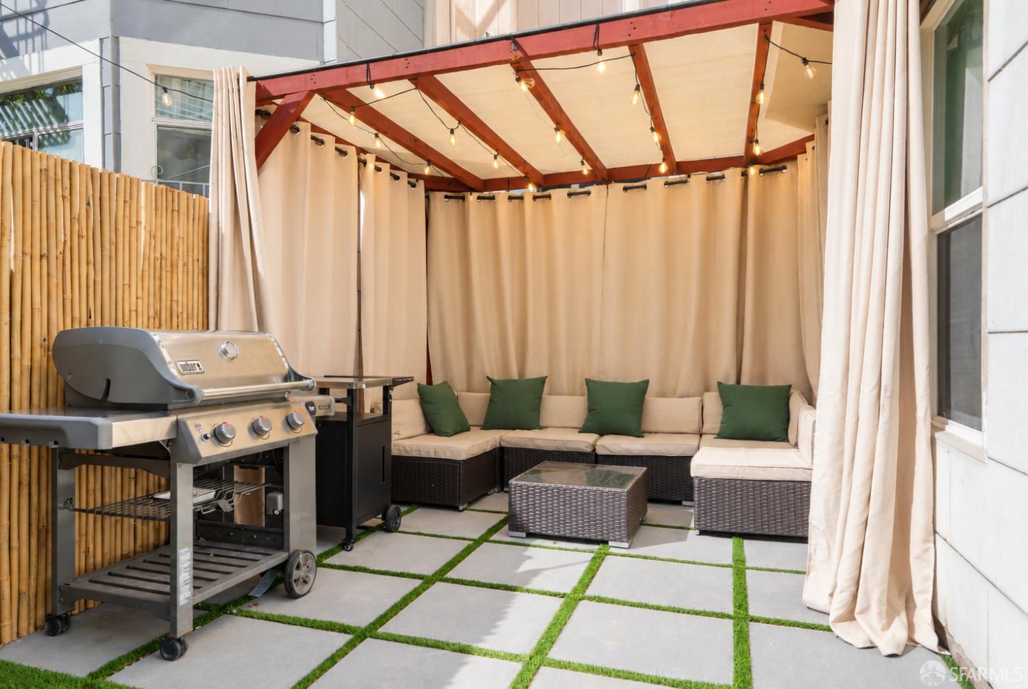 Covered patio with cushioned sectional seating, string lights, a gas grill, and grass between stone pavers.