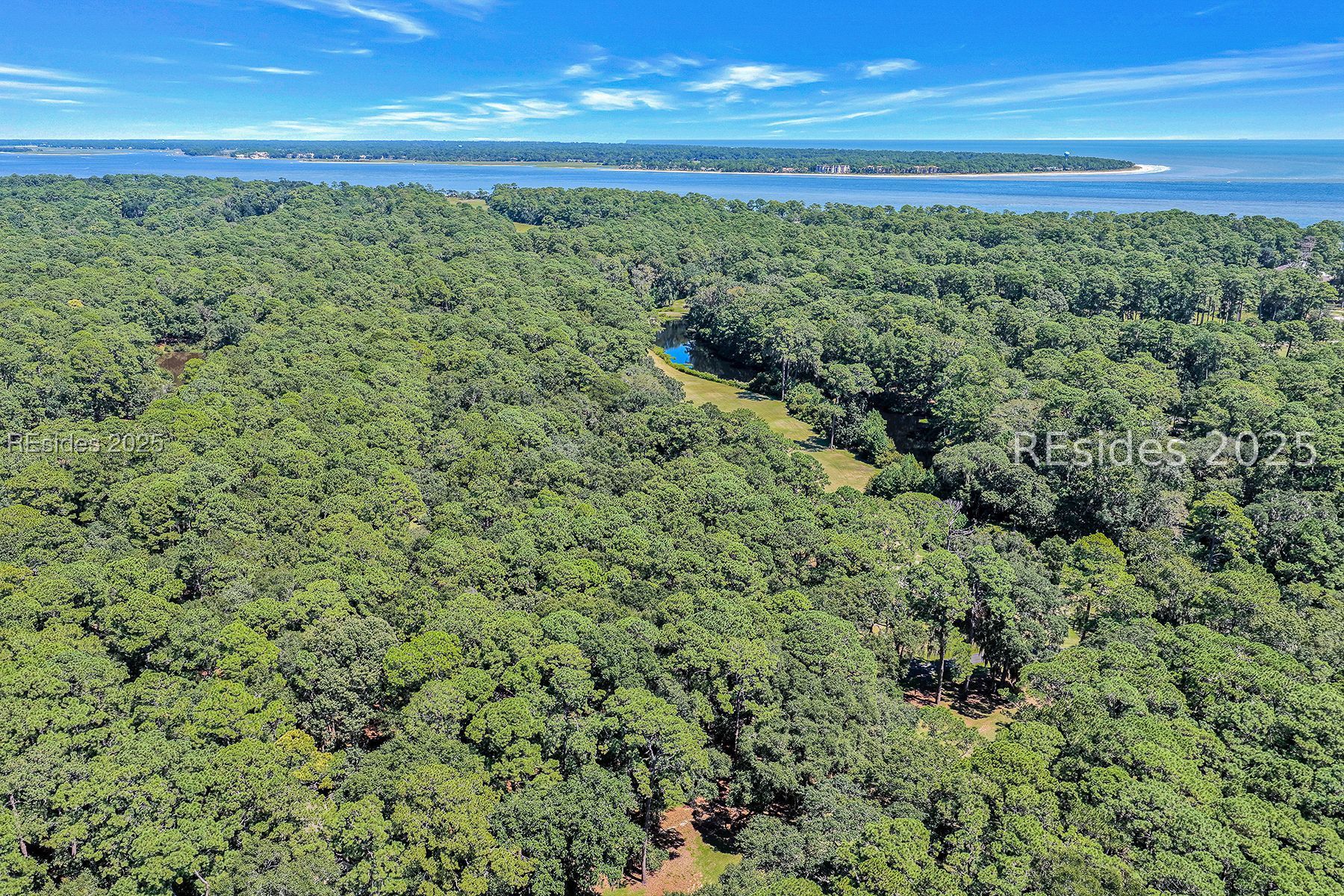 Aerial view of wooded land with a grassy clearing, waterway, and coastal shoreline beyond.