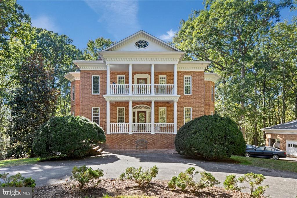 Red-brick house with white columns and two balconies, framed by mature trees.