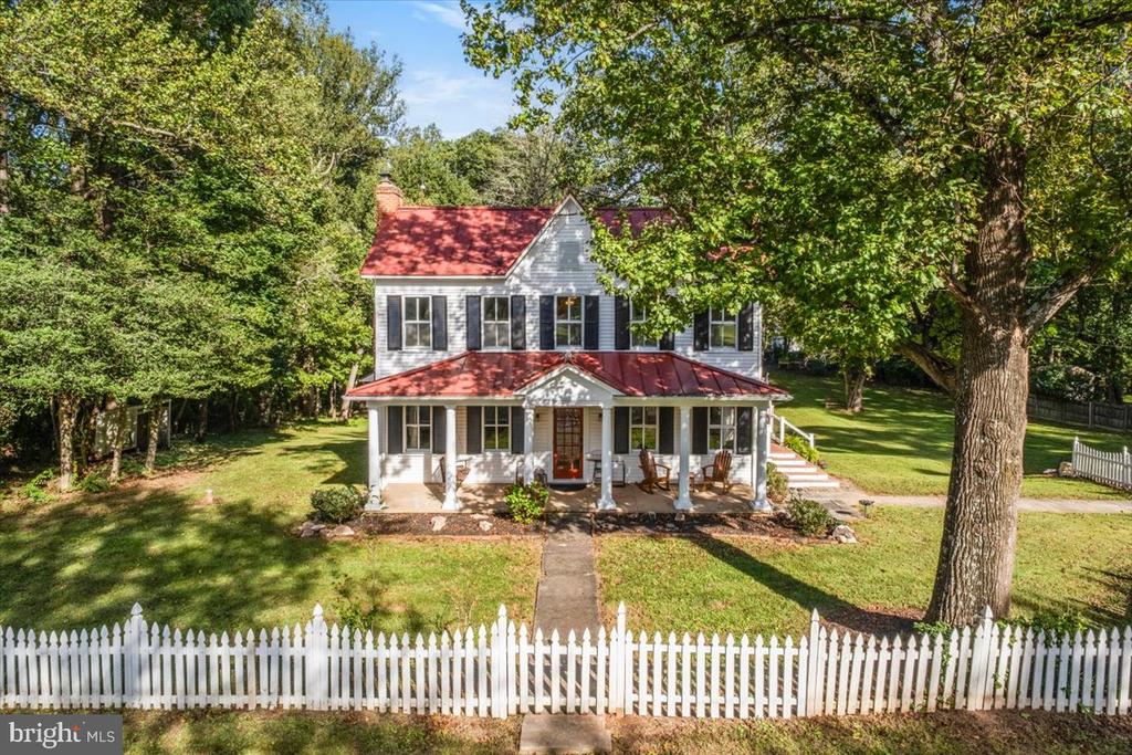 Two-story gray house with a red roof, covered porch, white picket fence, and mature trees.