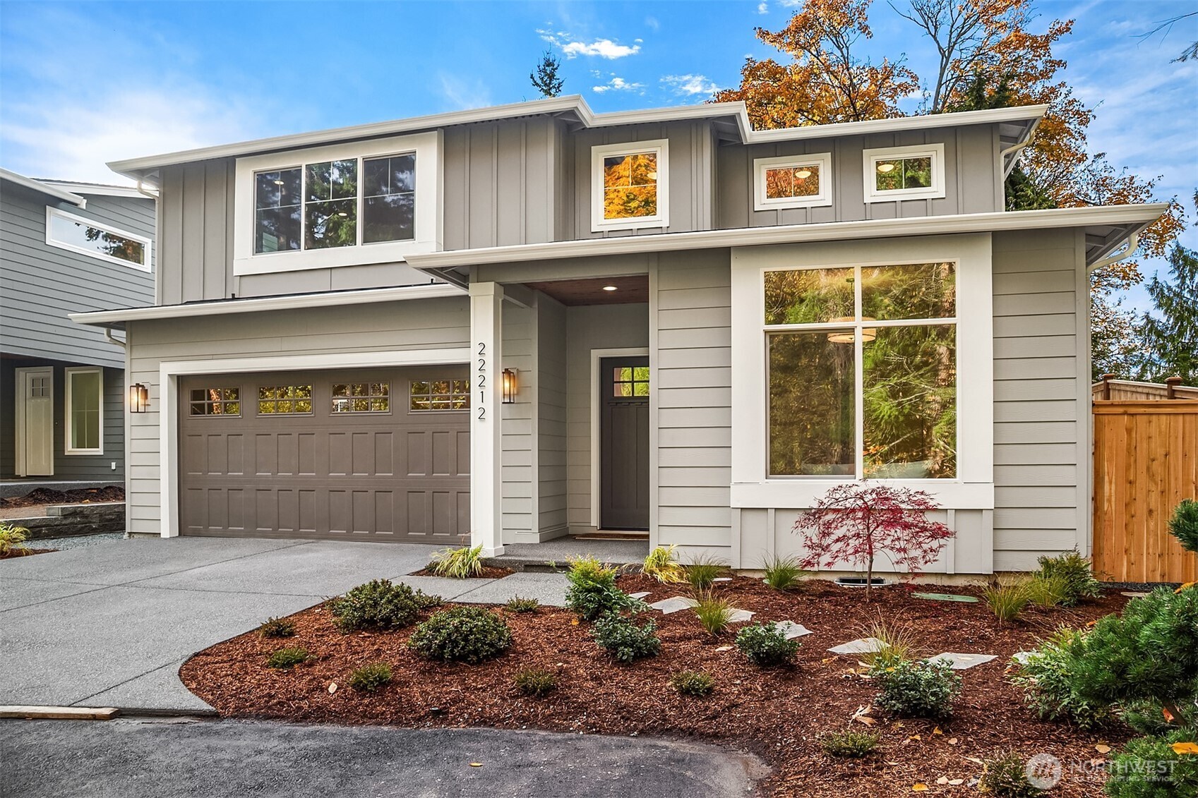 Modern gray two-story home with a double garage, landscaped beds, and autumn foliage.