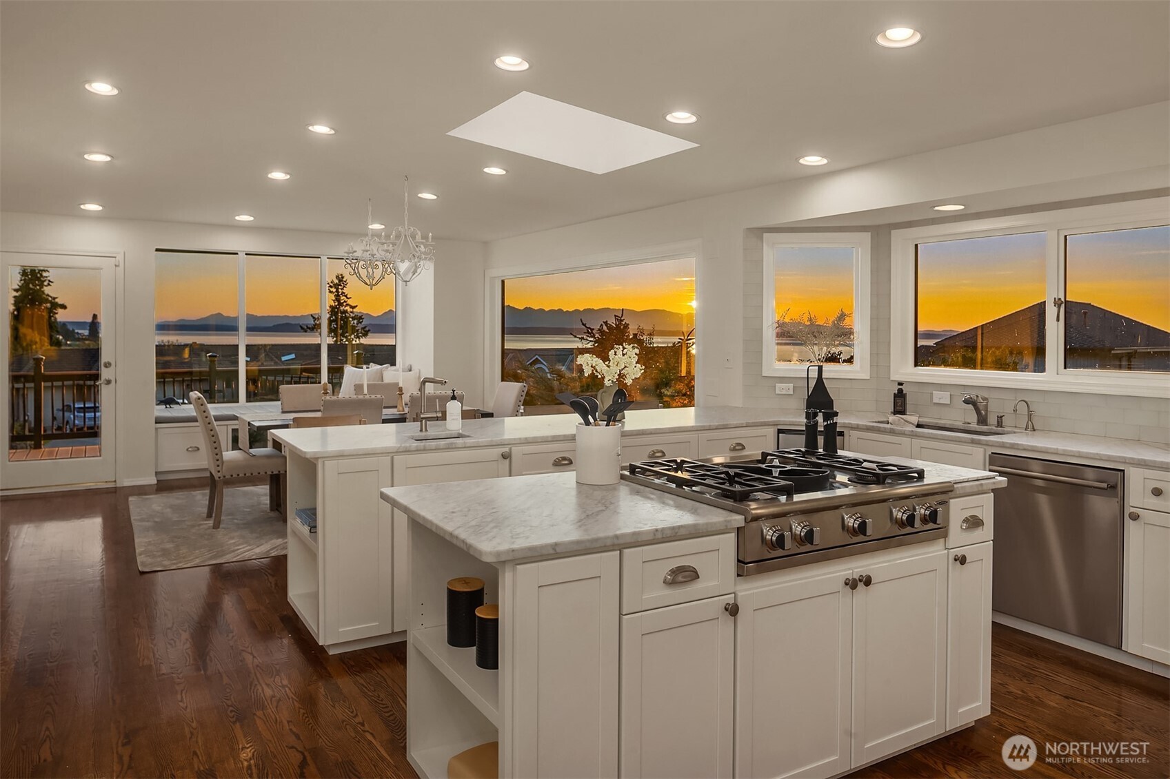 Furnished white kitchen with a marble island and sunset water and mountain views through large windows.