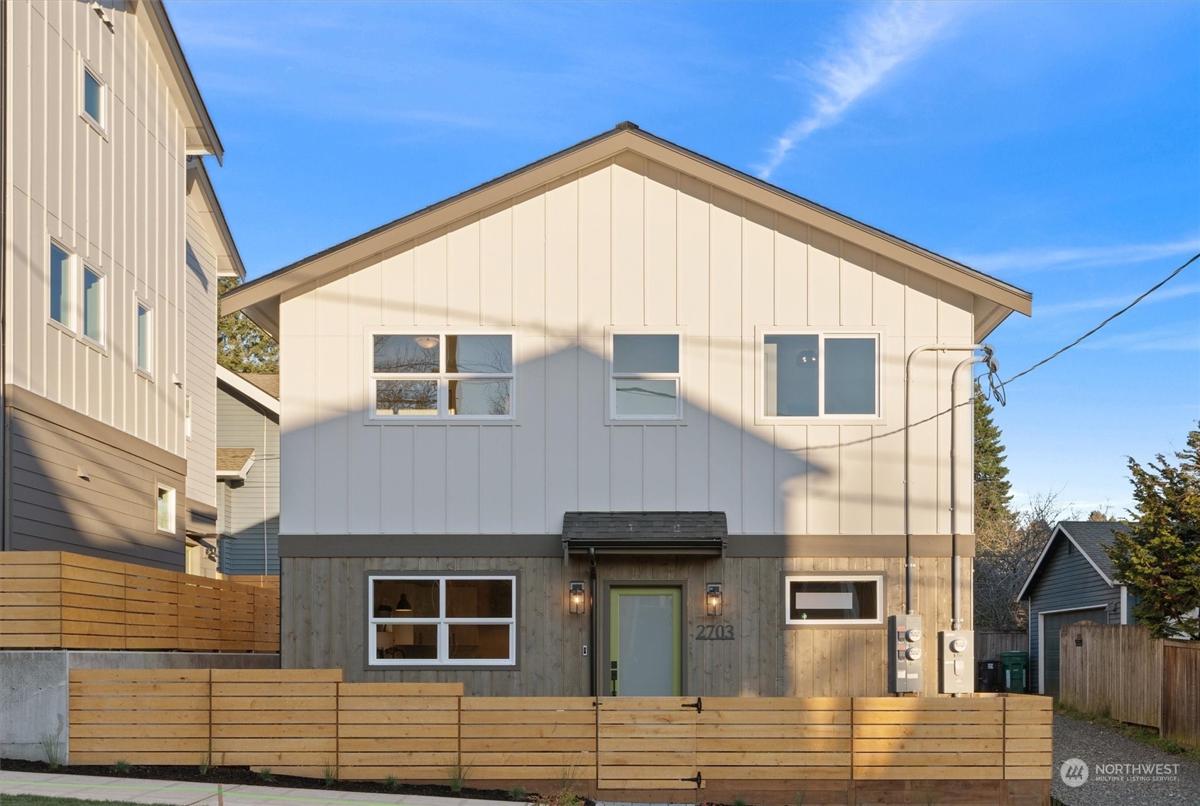 Modern two-story home with white siding, wood-clad lower level, green door, and horizontal wood fence.