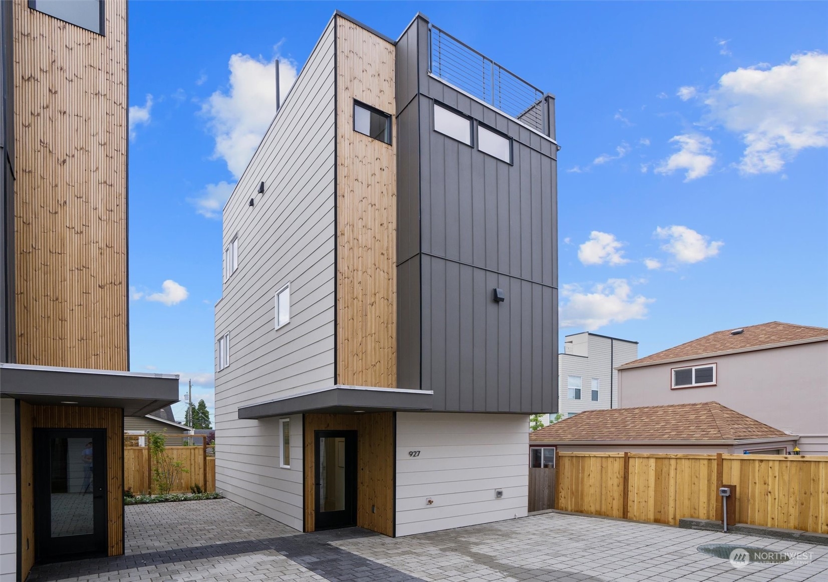 Modern narrow home with gray siding, wood accents, rooftop deck, and paved courtyard.