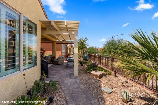 Backyard patio with a pergola-covered seating area, pavers, desert plants, and a metal fence.