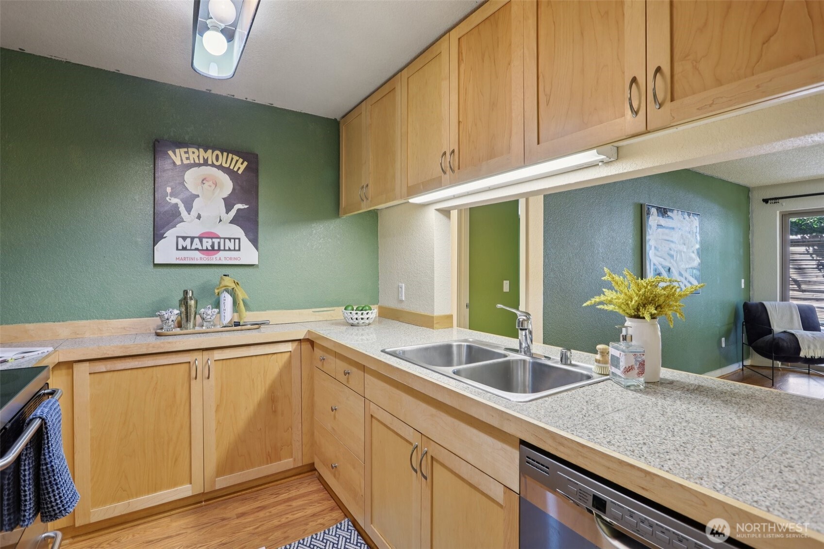 Kitchen with light wood cabinets, speckled countertops, and a double-basin sink; green walls and living area beyond.