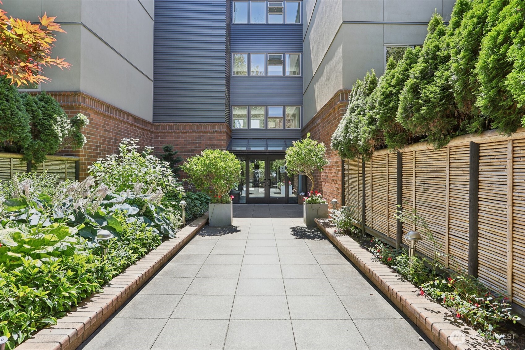 Residential building entrance at the end of a paved walkway, framed by landscaped beds and a slatted fence.
