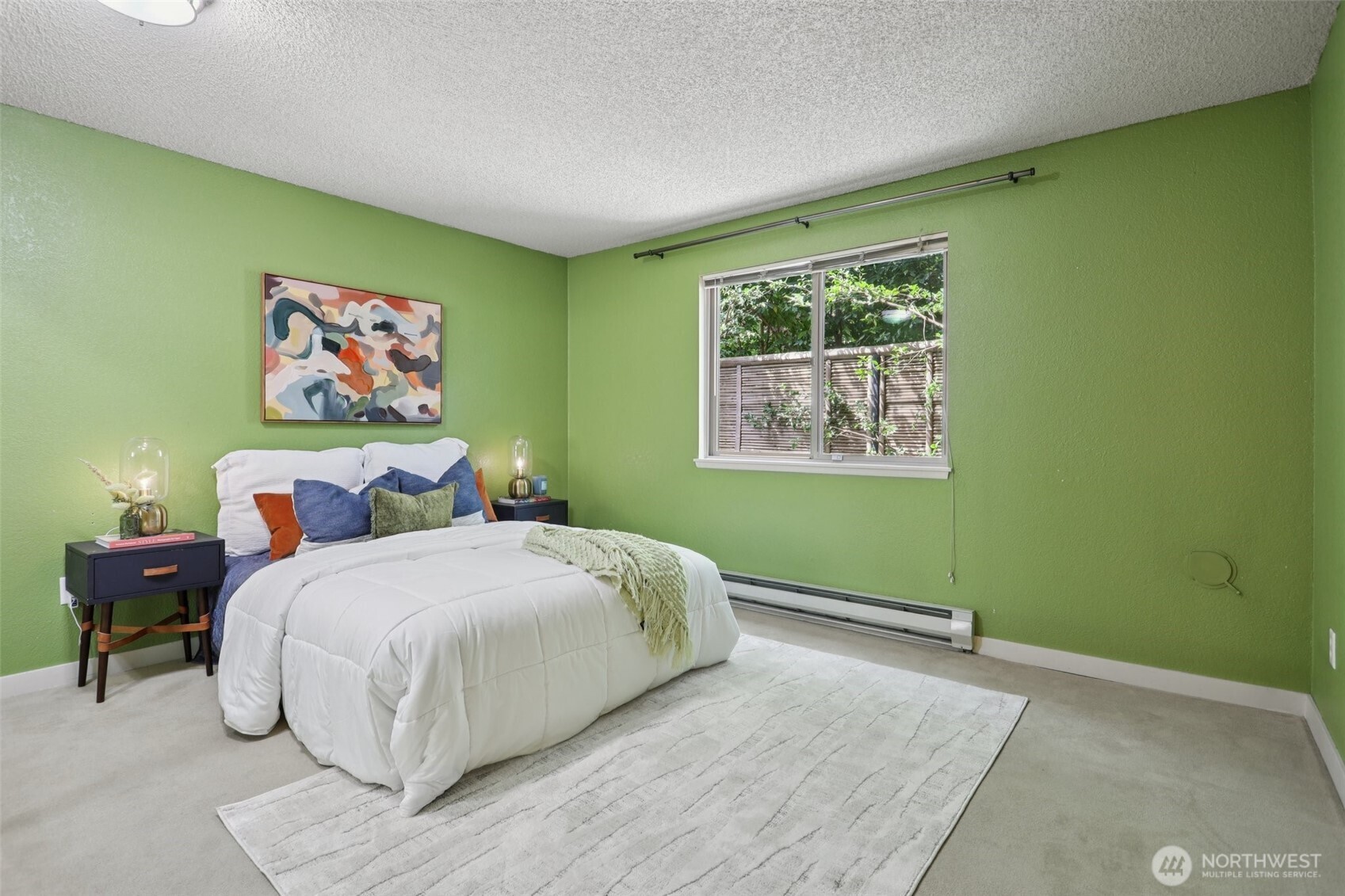 Staged bedroom with green walls, a white bed, colorful pillows, and a window facing leafy greenery.