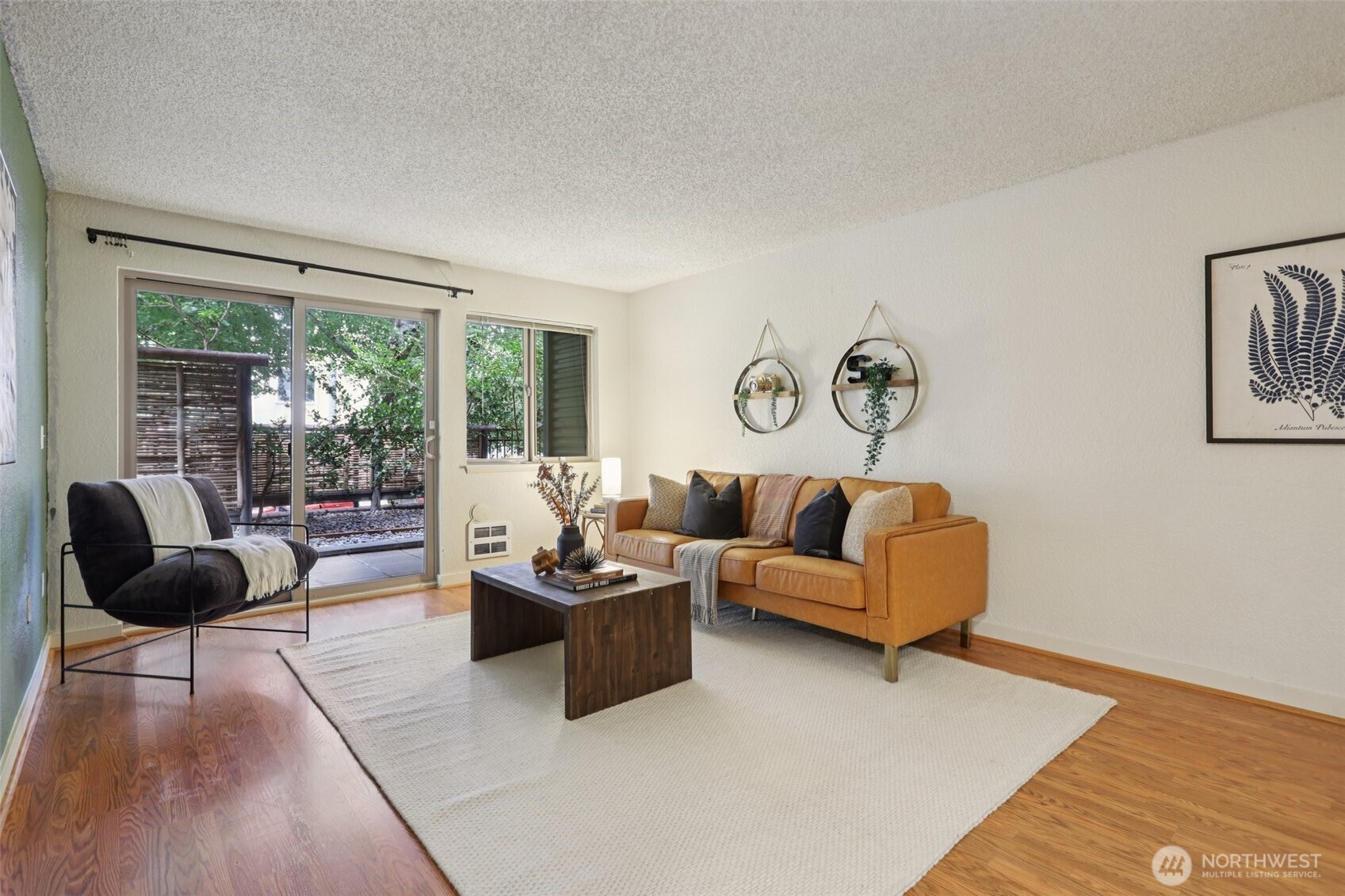 Living room with a tan leather sofa, dark chair, hardwood floors, and sliding doors to a leafy patio.