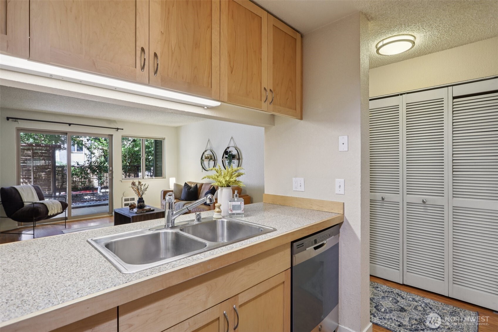 Kitchen with light wood cabinetry and double-basin sink, opening onto a living area and leafy patio.