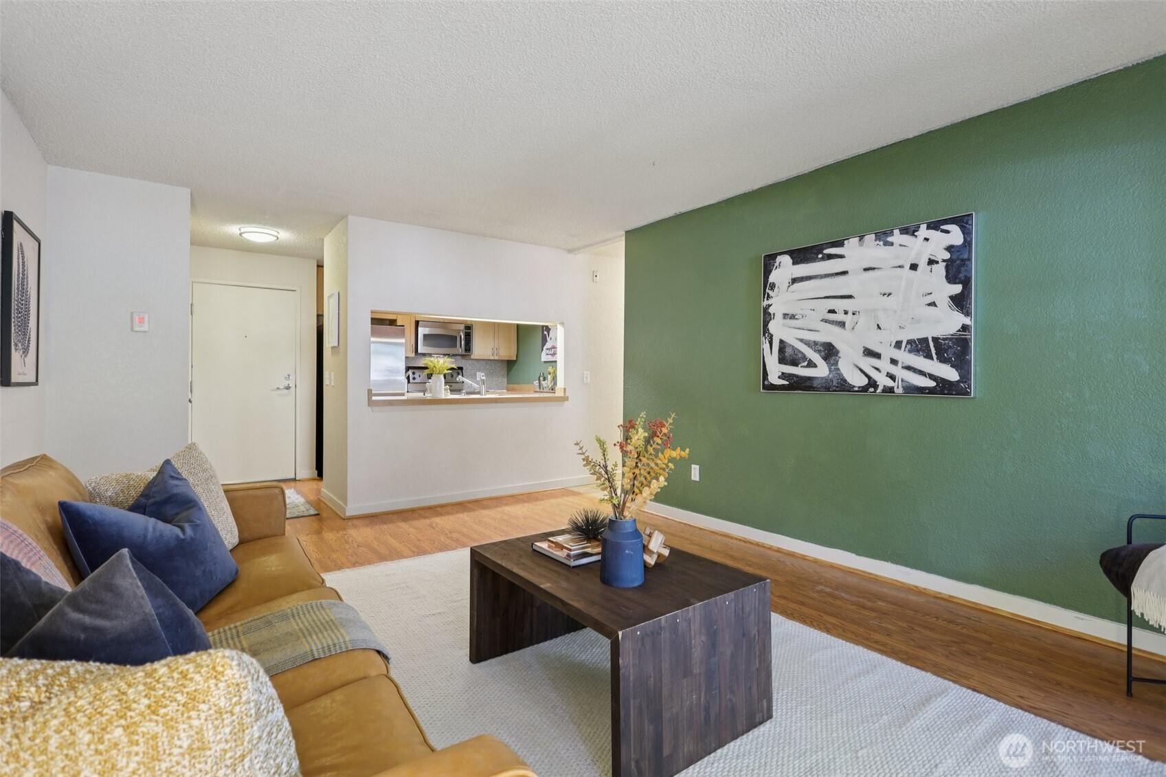Furnished living room with a tan sofa, green accent wall, and kitchen pass-through.