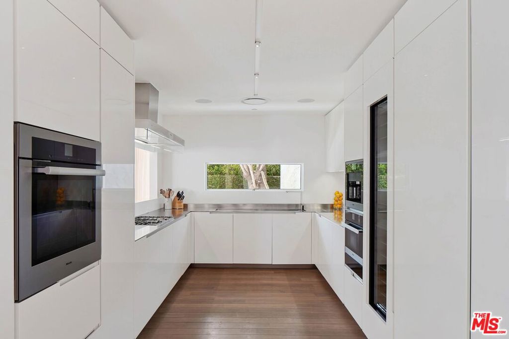 Modern white kitchen with built-in ovens, stainless steel counters, and a narrow window overlooking greenery.