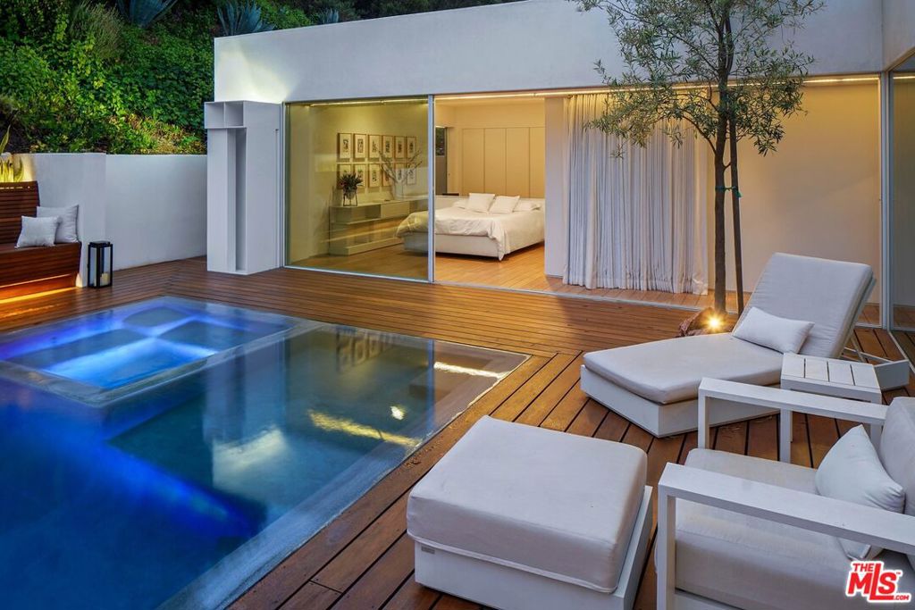 Modern home's illuminated pool and wood deck beside a glass-walled bedroom at dusk.