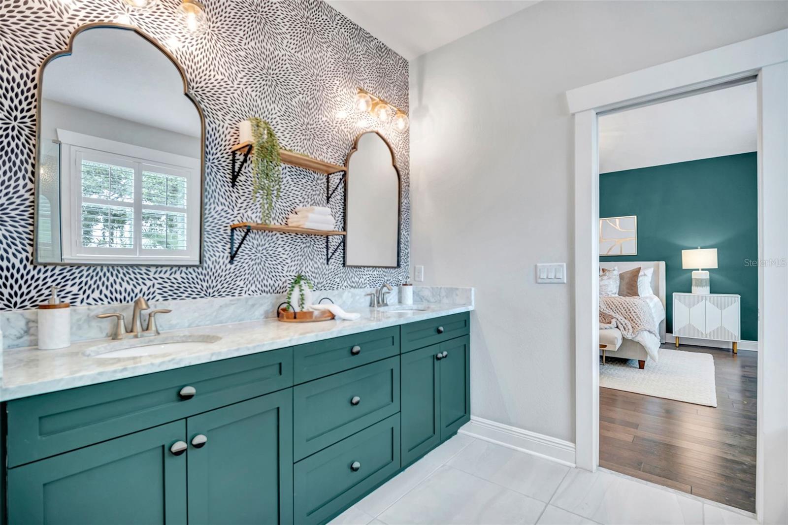 Bathroom with a teal double vanity, marble countertop, patterned wallpaper, and a view into a teal-accented bedroom.