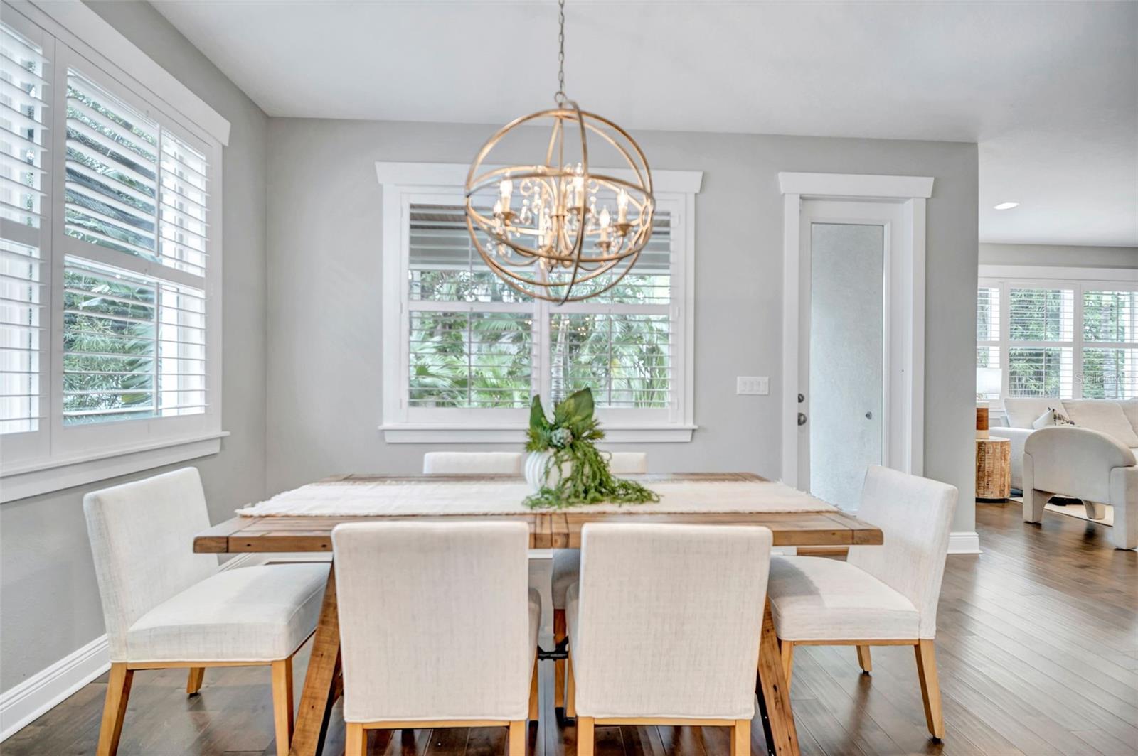 Dining room with a wooden table, white chairs, shuttered windows, and a brass globe chandelier.