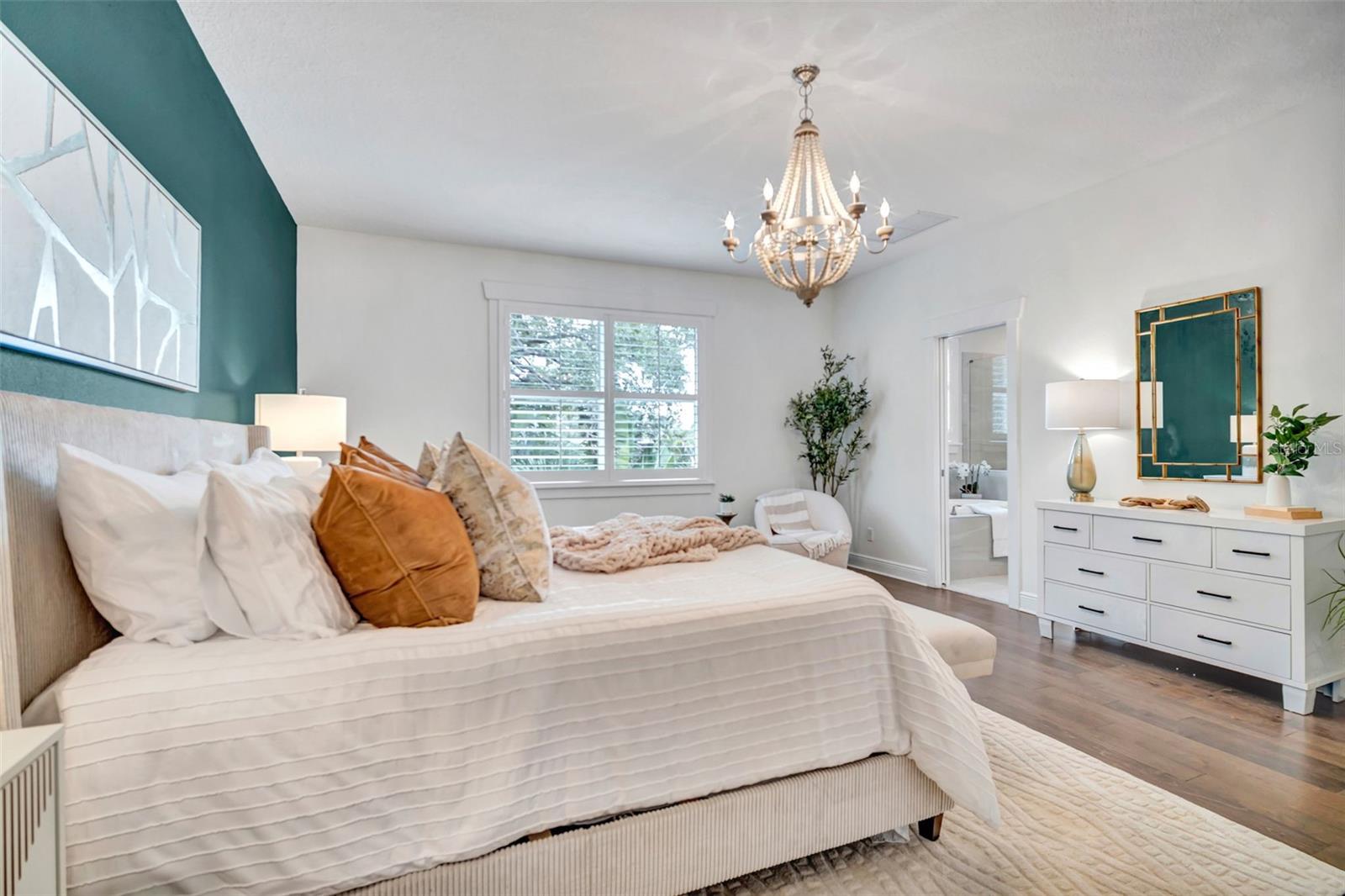 Staged bedroom with a white bed, teal accent wall, chandelier, and shuttered window.