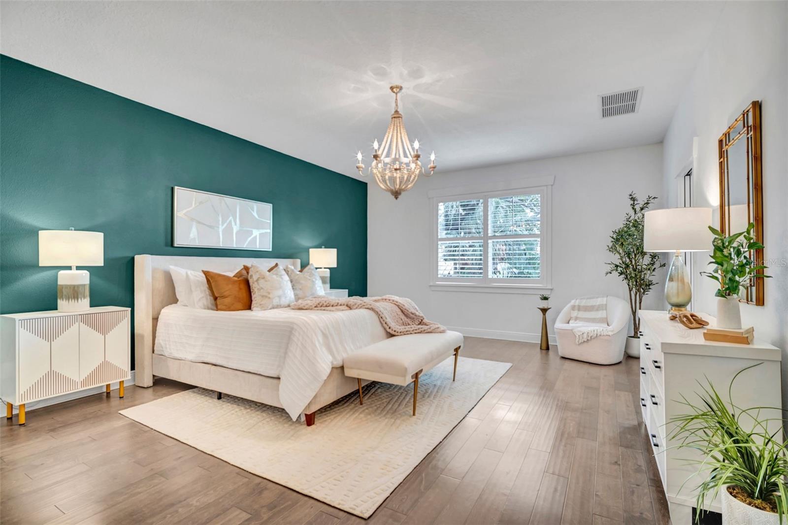 Staged bedroom with a white bed, teal accent wall, chandelier, and window seating area.