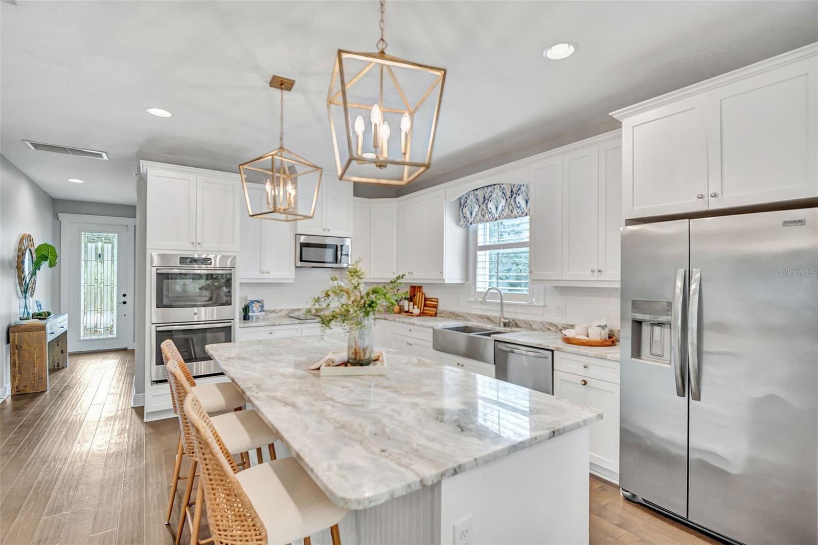 Bright white kitchen with a large stone island, stainless steel appliances, and brass pendant lights.