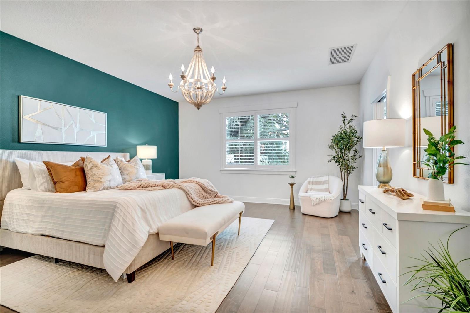 Furnished bedroom with a teal accent wall, white bed, chandelier, and gold-framed mirror.