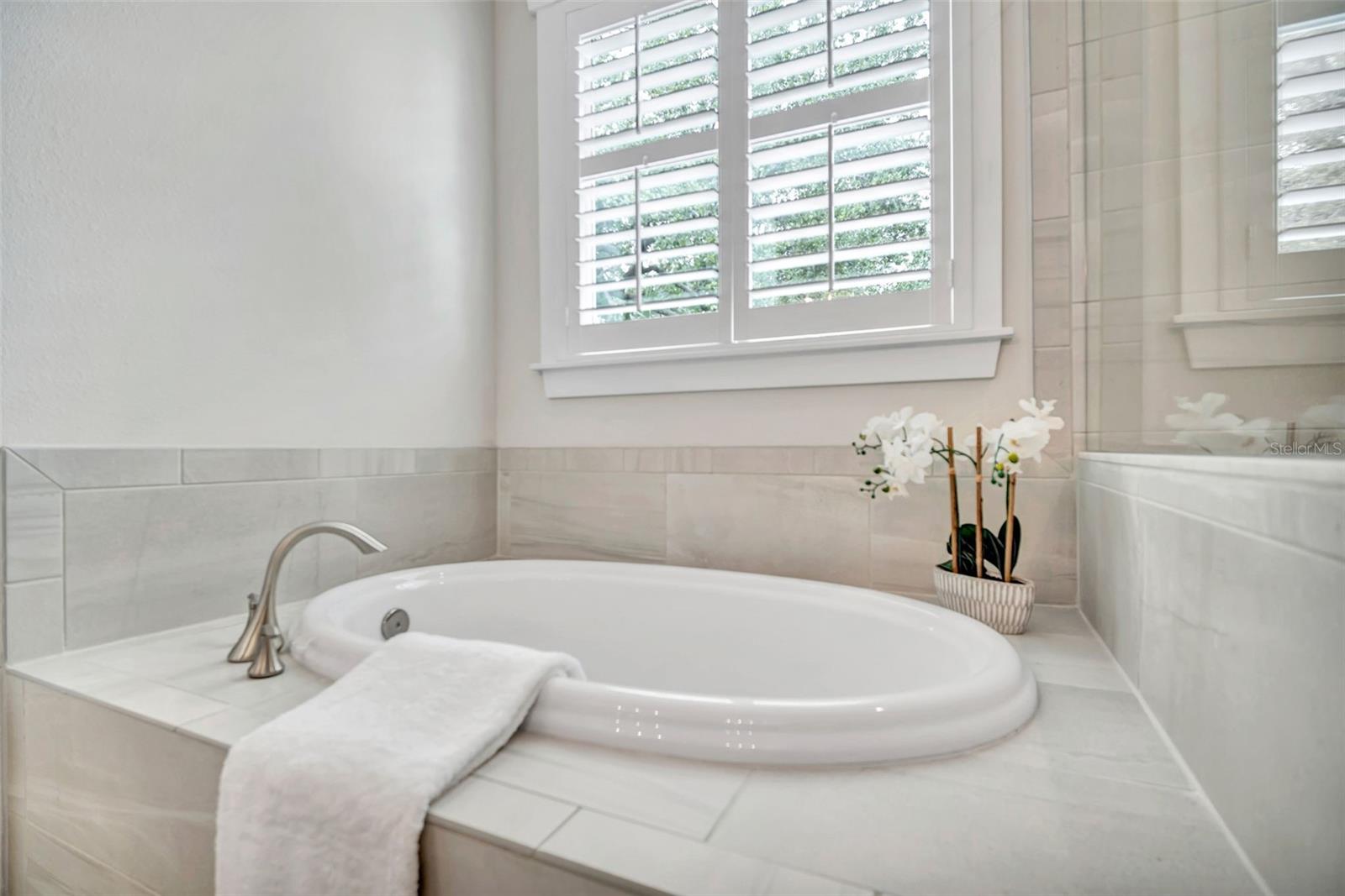 Bathroom soaking tub with pale tile, plantation shutters, and an orchid on the ledge.