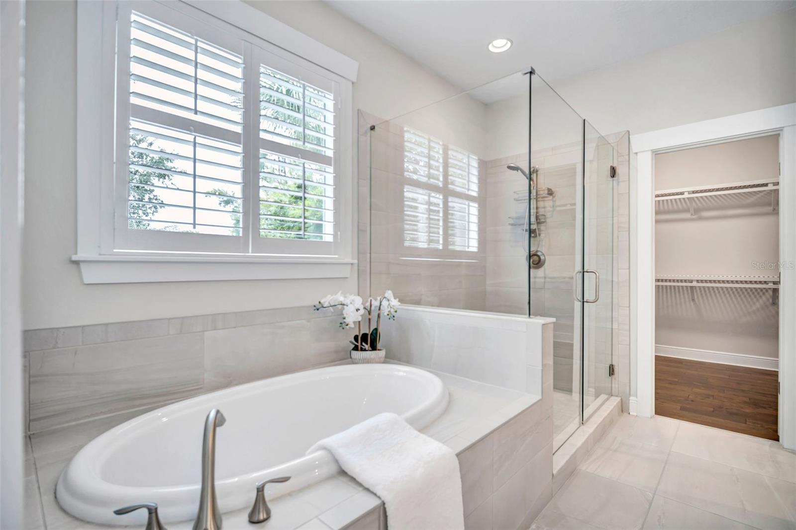 Primary bathroom with a soaking tub, glass shower, plantation shutters, and an open walk-in closet.