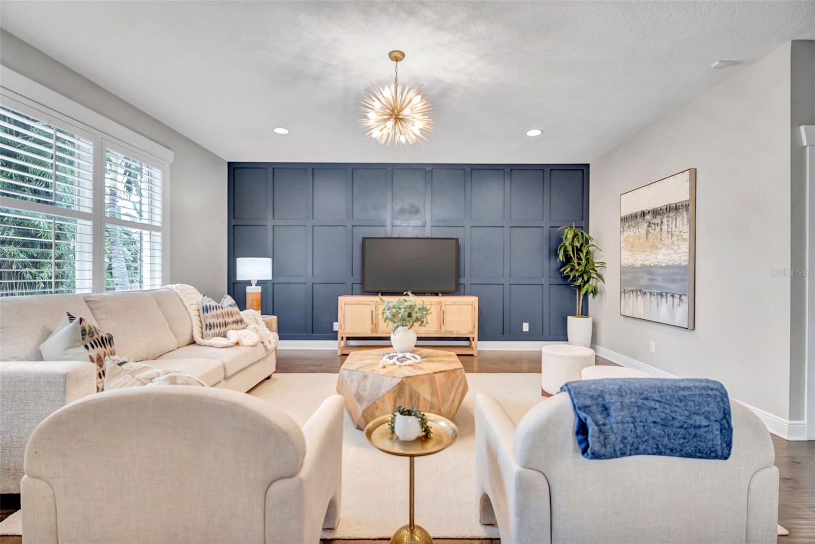 Staged living room with a dark blue paneled TV wall, beige seating, and shuttered windows.