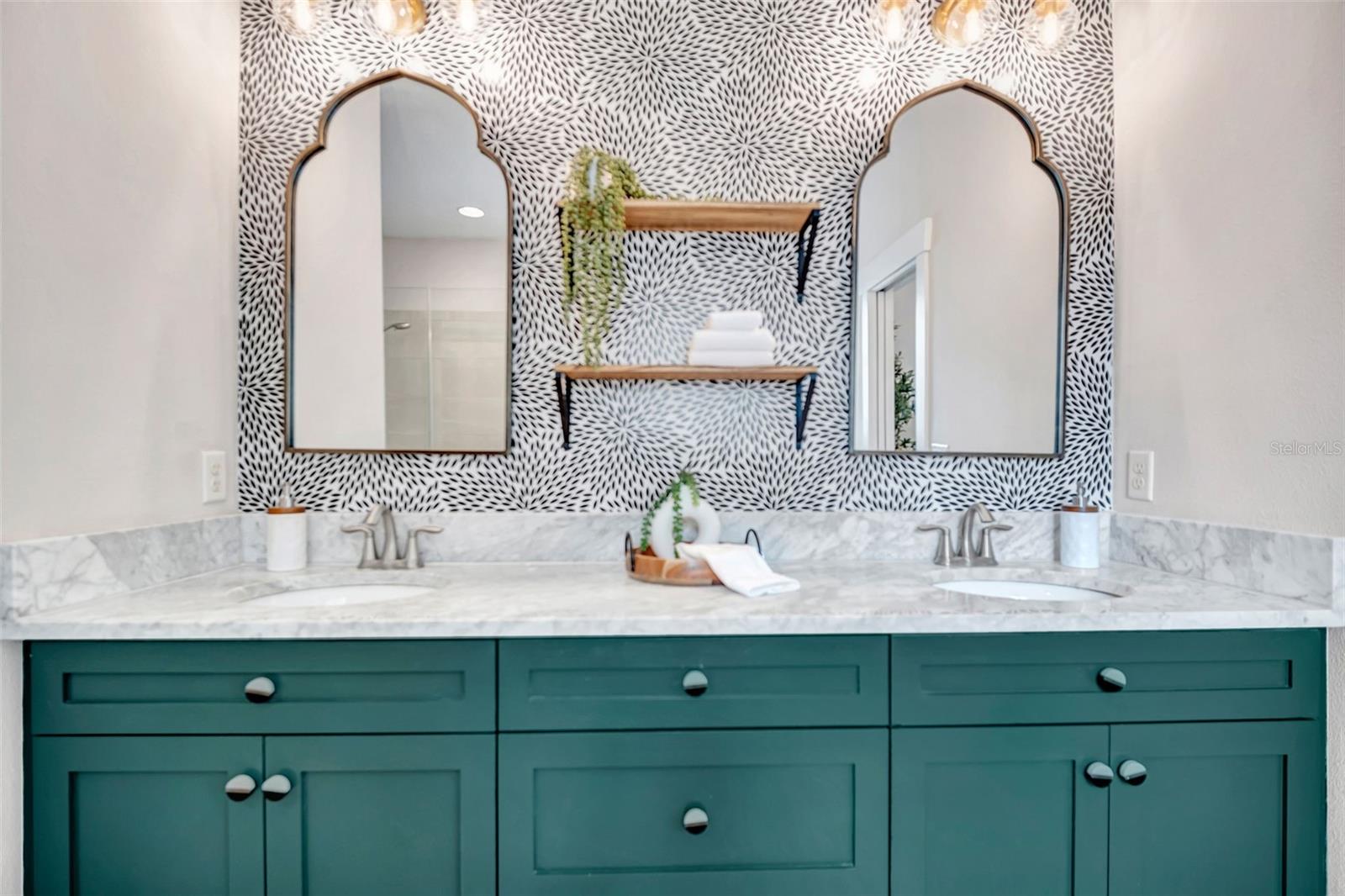 Bathroom vanity with teal cabinets, marble countertop, patterned wallpaper, and two arched mirrors.
