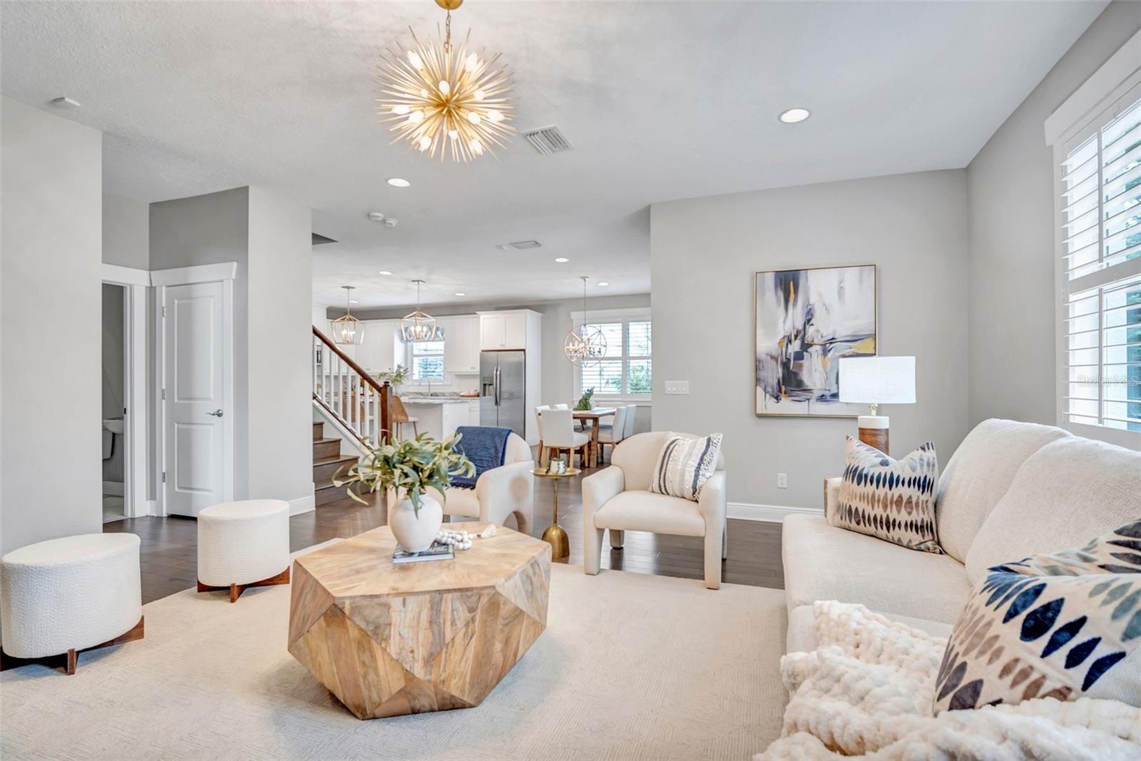 Staged living room opening to a white kitchen and dining area, with a geometric wood coffee table and starburst chandelier.