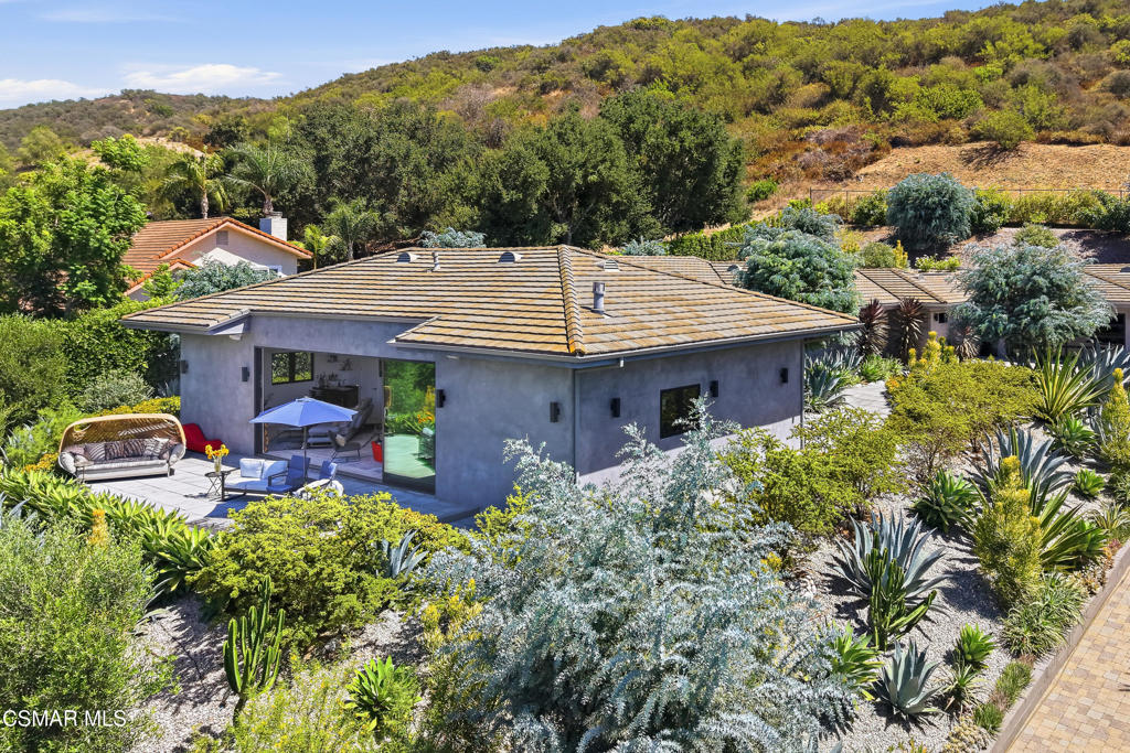 Single-story gray home with a tile roof, patio seating, drought-tolerant garden, and wooded hillside backdrop.