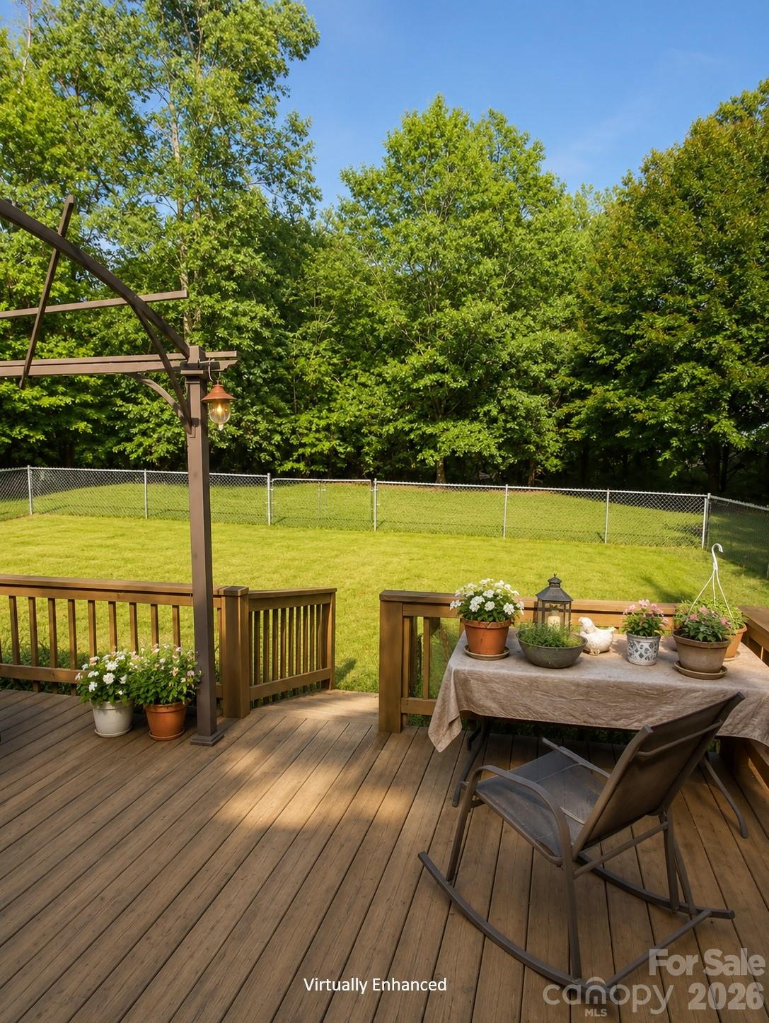 Wooden deck with a pergola and flower-filled dining table overlooking a fenced, tree-lined lawn.