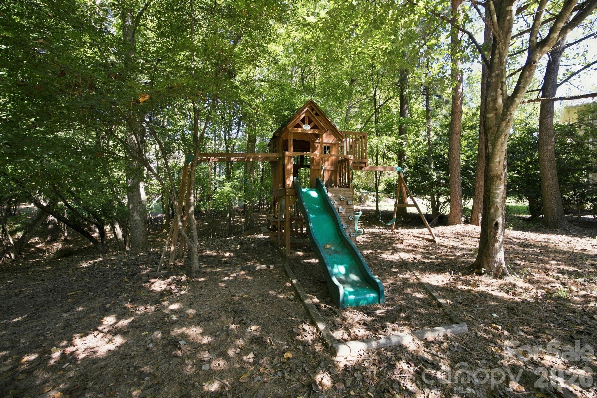 Backyard playset with a wooden treehouse, green slide, climbing wall, and swings beneath mature trees.