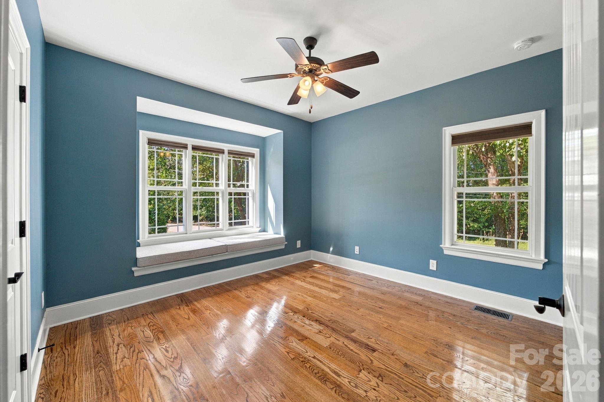 Vacant blue room with polished hardwood floors, a built-in window seat, and a ceiling fan.