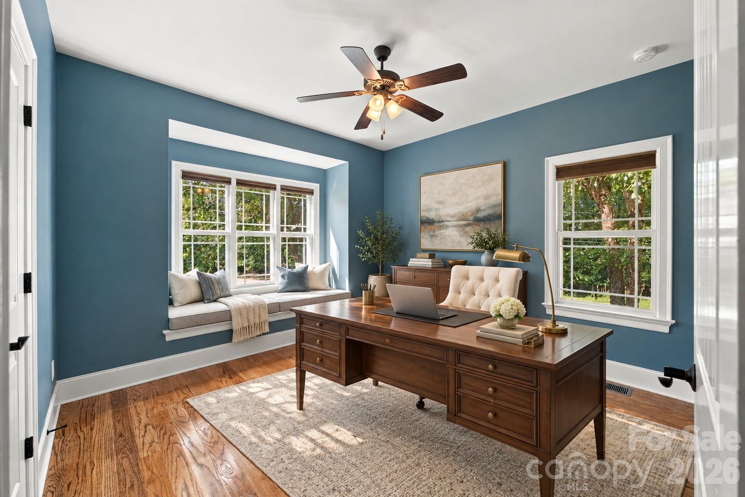 Blue home office with a large wooden desk and a cushioned window seat beneath three windows.