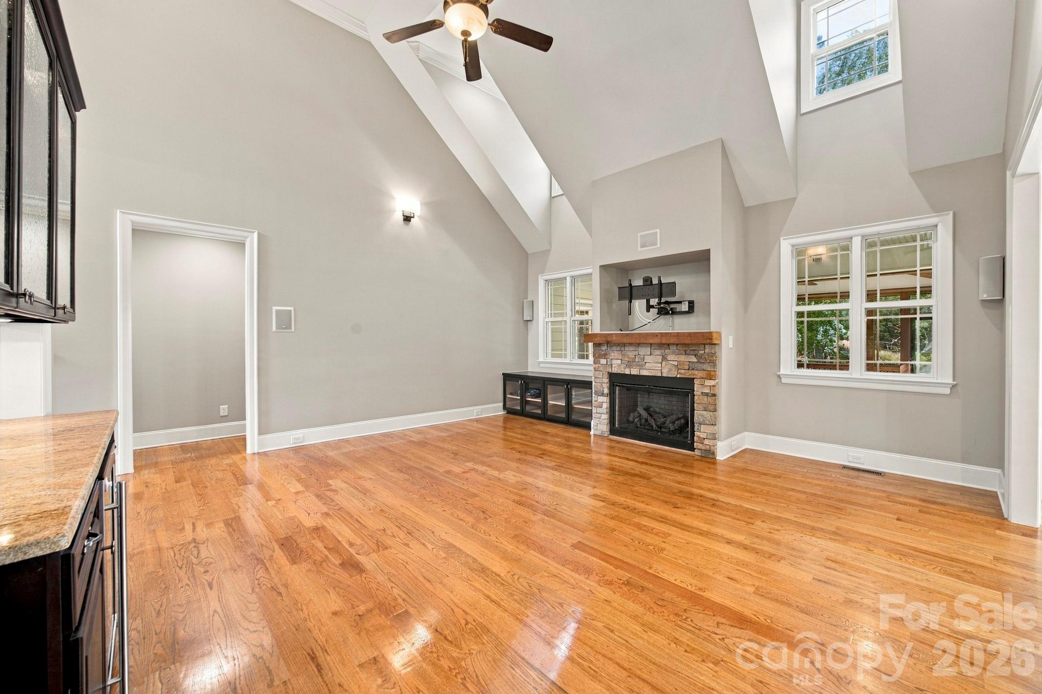Vacant living room with vaulted ceiling, polished hardwood floors, and a stone fireplace.