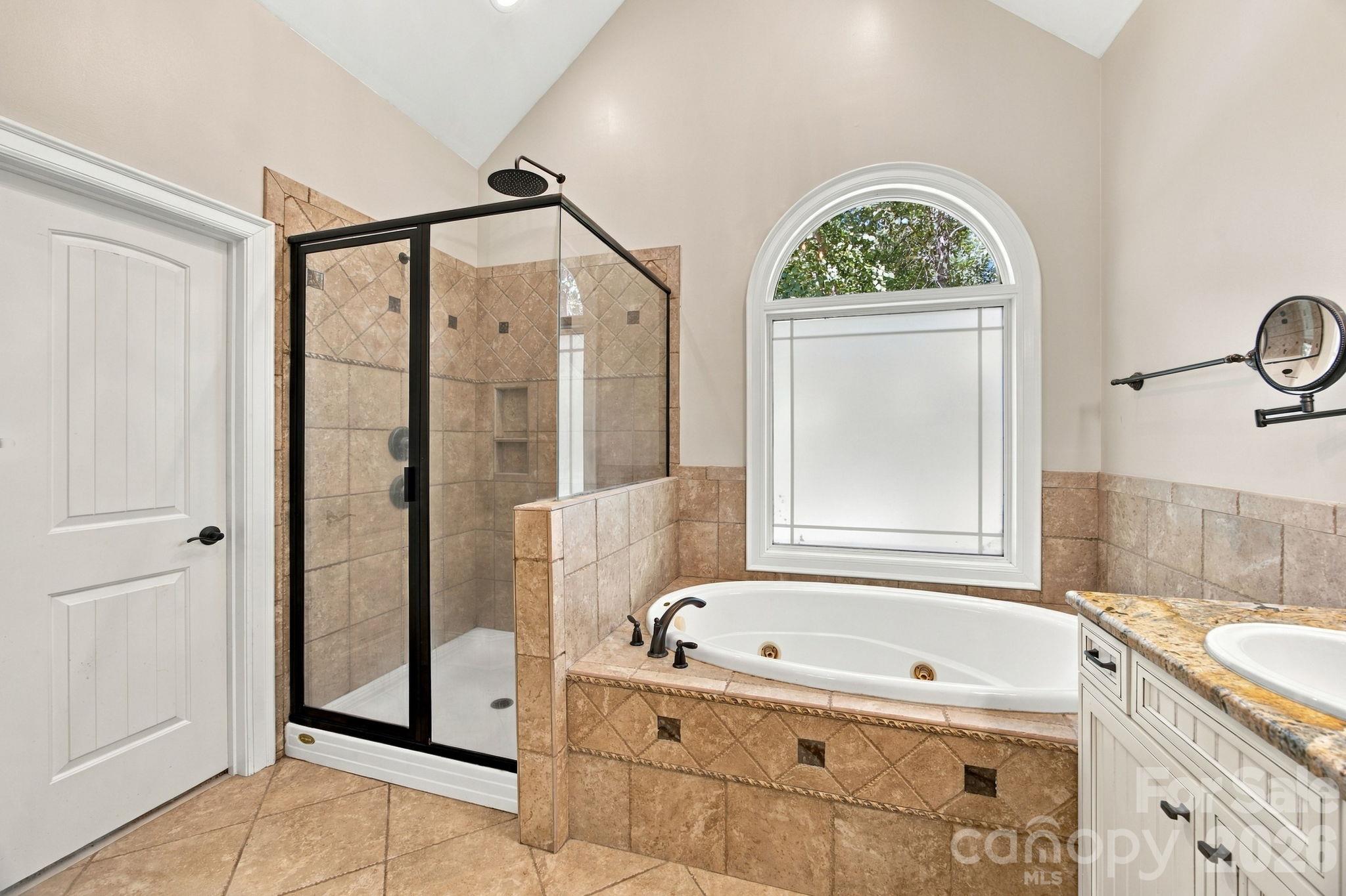 Bathroom with a glass walk-in shower, tiled soaking tub, arched window, and granite vanity.